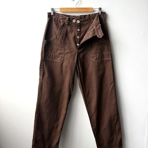 May include: Dark brown pants featuring a high-waisted design with a button-fly closure. These pants include two large front patch pockets and are displayed on a wooden hanger, showcasing a straight-leg cut and visible stitching details throughout.