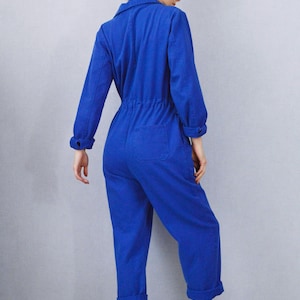 May include: A vibrant blue jumpsuit with long sleeves, a collared neckline, and a cinched waist. The jumpsuit features a back pocket and is paired with white high-top sneakers. The cuffs of the sleeves and pants are rolled up.