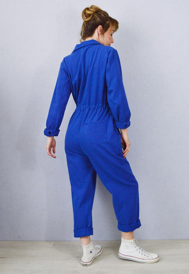 May include: A vibrant blue jumpsuit with long sleeves, a collared neckline, and a cinched waist. The jumpsuit features a back pocket and is paired with white high-top sneakers. The cuffs of the sleeves and pants are rolled up.
