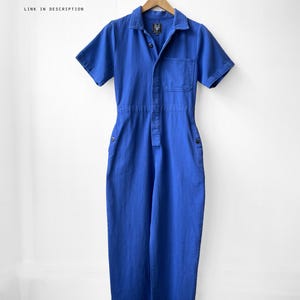 May include: A short-sleeved, blue jumpsuit hangs on a wooden hanger. The jumpsuit has a collared neckline, a front pocket, and button closures. The text "SHORT SLEEVED VERSION AVAILABLE LINK IN DESCRIPTION" is visible in the image.
