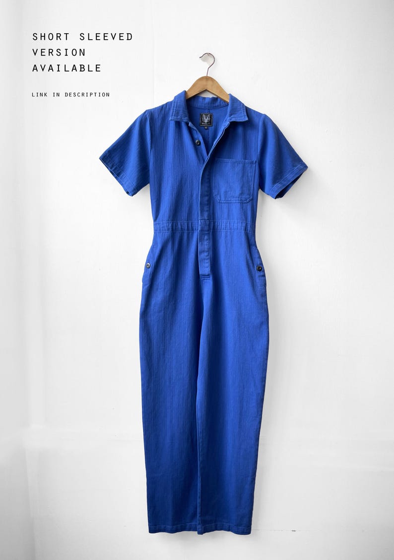 May include: A short-sleeved, blue jumpsuit hangs on a wooden hanger. The jumpsuit has a collared neckline, a front pocket, and button closures. The text "SHORT SLEEVED VERSION AVAILABLE LINK IN DESCRIPTION" is visible in the image.