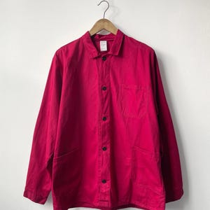 May include: A bright red work jacket with a collared neckline, long sleeves, and black buttons. The jacket has two front pockets and is hanging on a wooden hanger. The garment is made of a sturdy fabric.