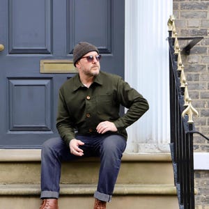 May include: A dark olive green button-down shirt jacket with a chest pocket, dark gray denim jeans with cuffed hems, brown leather lace-up boots, a dark brown knit beanie, and brown-tinted sunglasses. The outfit is worn as a complete ensemble.