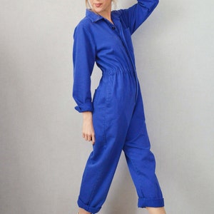 May include: A cobalt blue jumpsuit with long sleeves, a collared neckline, and a button-down front. The jumpsuit features a cinched waist and rolled-up cuffs. The model is wearing white high-top sneakers.