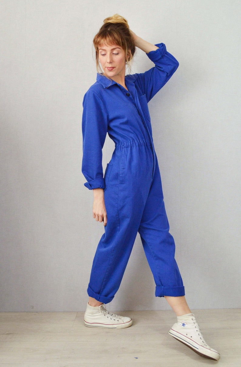 May include: A cobalt blue jumpsuit with long sleeves, a collared neckline, and a button-down front. The jumpsuit features a cinched waist and rolled-up cuffs. The model is wearing white high-top sneakers.