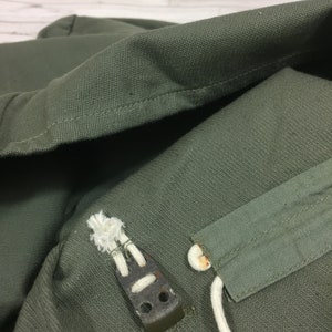 May include: Close-up of a green canvas bag with a white drawstring and a metal buckle. The bag is made of a durable material and has a simple design.