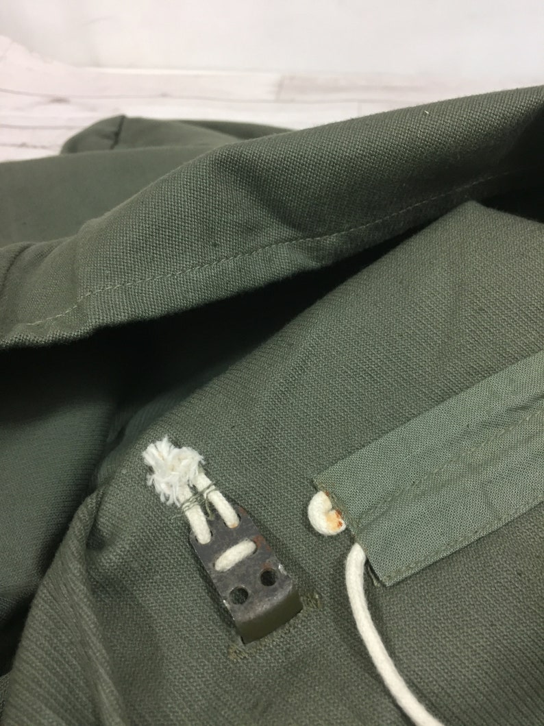 May include: Close-up of a green canvas bag with a white drawstring and a metal buckle. The bag is made of a durable material and has a simple design.