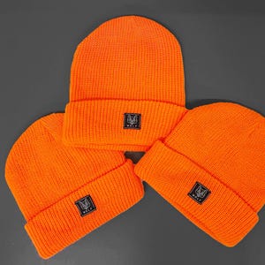 Peut inclure: Trois bonnets en tricot orange vif avec un patch logo de loup noir. Les bonnets sont pliés et empilés sur une surface grise.