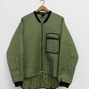 May include: An olive green quilted jacket with black trim and a zip closure. The jacket features a black pocket and is hanging on a wooden hanger. The jacket has a quilted design and a bomber-style collar.