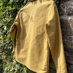 May include: Mustard-colored long-sleeve shirt with a button-down front and collar. The shirt is made of a soft, textured fabric and has buttoned cuffs. The garment is displayed against a stone wall and green foliage.