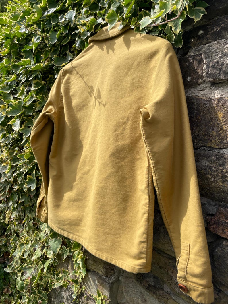 May include: Mustard-colored long-sleeve shirt with a button-down front and collar. The shirt is made of a soft, textured fabric and has buttoned cuffs. The garment is displayed against a stone wall and green foliage.