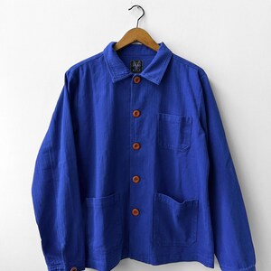 May include: A vibrant blue long-sleeve jacket featuring a classic collar, a front button closure with five brown buttons, one chest pocket, and two lower patch pockets. The interior neck label displays the text WOLF.