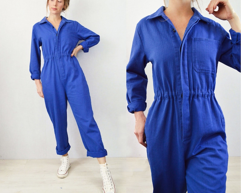 May include: A vibrant blue jumpsuit with long sleeves, a collared neckline, and a front pocket. The waist is cinched with an elastic band. The pants are cuffed at the ankles. The model is wearing white sneakers.