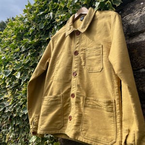 May include: Mustard-colored jacket with a button-up front, two chest pockets, and two lower pockets. The jacket has a classic collar and brown buttons. It is hanging on a wooden hanger against a backdrop of ivy and a stone wall.