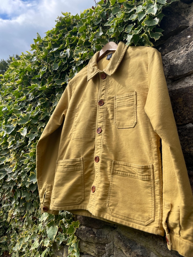 May include: Mustard-colored jacket with a button-up front, two chest pockets, and two lower pockets. The jacket has a classic collar and brown buttons. It is hanging on a wooden hanger against a backdrop of ivy and a stone wall.