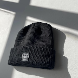 May include: Black knit beanie with a folded brim. A white square patch with a wolf graphic and the word "WOLF" is sewn on the front. The hat is made of a ribbed knit material.