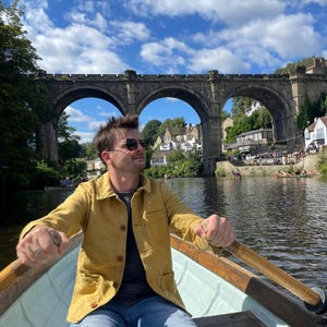 May include: A person rowing a boat on a river under a stone bridge with multiple arches. The person is wearing a yellow jacket, blue jeans, and sunglasses. The sky is blue with fluffy white clouds.