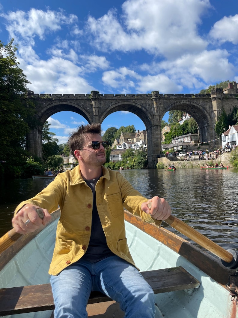 May include: A person rowing a boat on a river under a stone bridge with multiple arches. The person is wearing a yellow jacket, blue jeans, and sunglasses. The sky is blue with fluffy white clouds.