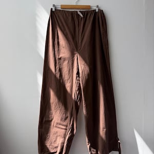 May include: Wide-leg trousers in a solid dark brown colour featuring an elasticated waistband with a drawstring closure. The trousers have a relaxed, loose fit throughout the legs and include adjustable tie details at the bottom of each hem.