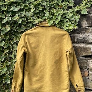 May include: Mustard-colored jacket with a button-down front, long sleeves, and a collared neckline. The jacket is made of a sturdy fabric and has two buttoned pockets. The jacket is hanging against a stone wall and green foliage.