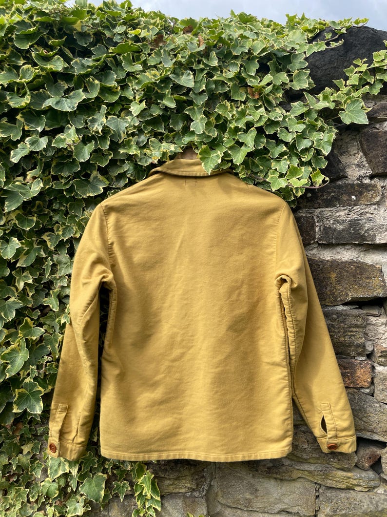 May include: Mustard-colored jacket with a button-down front, long sleeves, and a collared neckline. The jacket is made of a sturdy fabric and has two buttoned pockets. The jacket is hanging against a stone wall and green foliage.