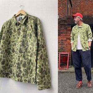 May include: A green and brown camouflage jacket with a button-up front and two chest pockets hangs on a wooden hanger. The jacket is made of a durable fabric and has a classic design. The jacket is shown on a white wall.
