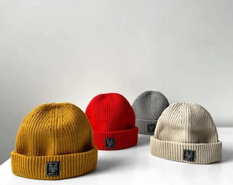 May include: Four ribbed knit beanies in mustard yellow, red, grey, and beige. Each beanie has a folded brim and a small black square patch with a wolf logo. The beanies are arranged on a white surface.