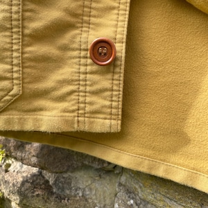 May include: Close-up of a mustard-colored jacket with a brown button. The jacket has vertical stitching details and a soft, textured interior. The button is round and has a four-hole design.