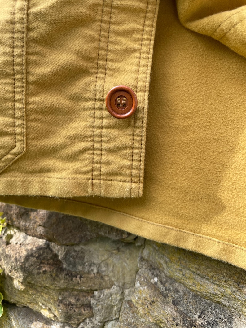 May include: Close-up of a mustard-colored jacket with a brown button. The jacket has vertical stitching details and a soft, textured interior. The button is round and has a four-hole design.