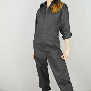May include: Dark grey jumpsuit featuring a collared neckline, long sleeves, and several pockets. The outfit is completed with a black cap and white trainers. The style is casual and practical.