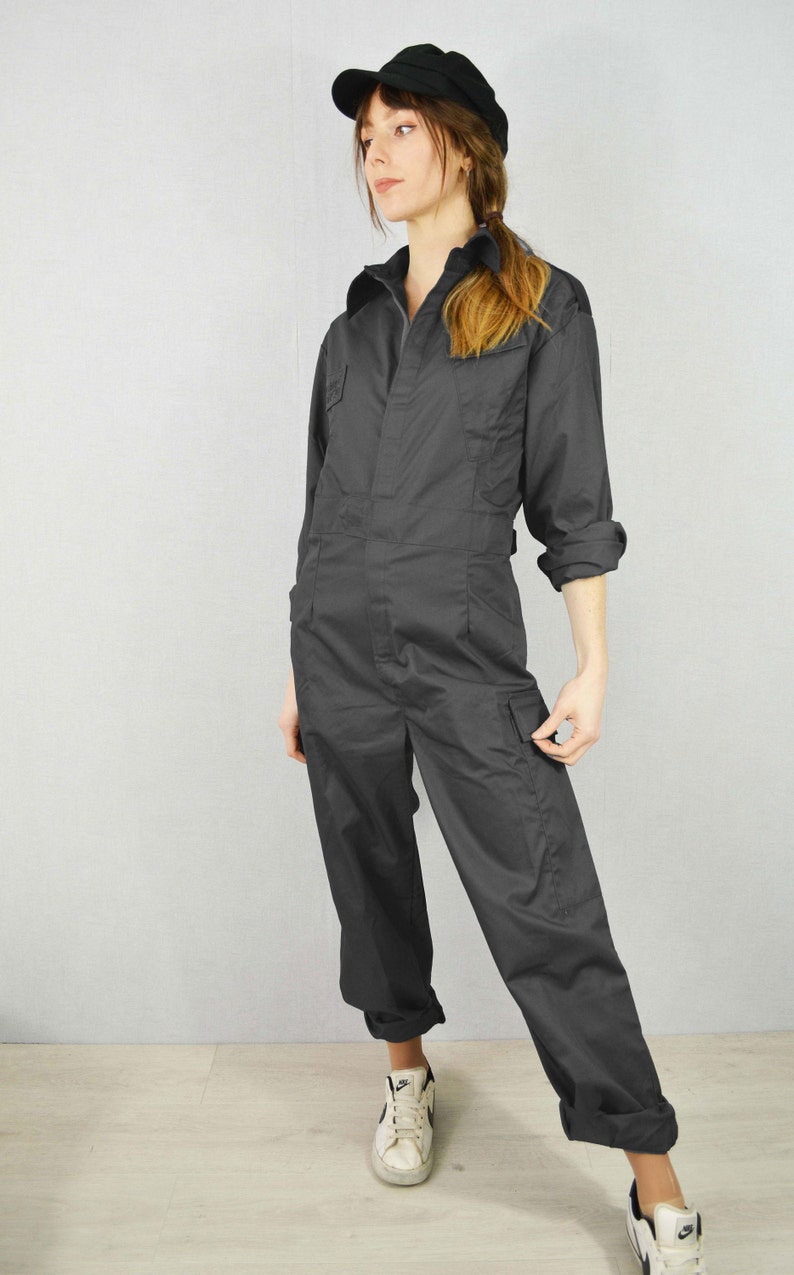 May include: Dark grey jumpsuit featuring a collared neckline, long sleeves, and several pockets. The outfit is completed with a black cap and white trainers. The style is casual and practical.