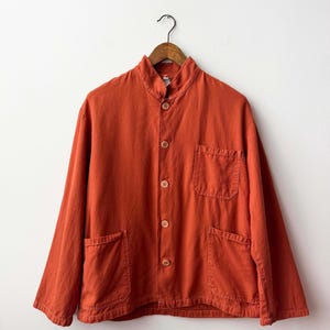 Peut inclure: Une veste orange brûlé à manches longues dotée d'un col officier, d'une fermeture à cinq boutons marron clair, d'une poche poitrine sur le côté gauche et de deux poches plaquées inférieures. La veste est présentée sur un cintre en bois.