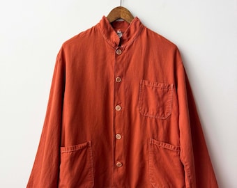 May include: A long-sleeved, burnt orange jacket featuring a mandarin collar, a front button closure with five light brown buttons, one chest pocket on the left side, and two lower patch pockets. The jacket is displayed on a wooden hanger.