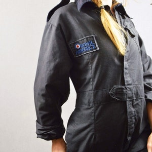 May include: Dark navy blue jumpsuit with a collared neckline and long sleeves. Features a belt at the waist and a patch on the chest that reads "ROYAL AIRFORCE" in blue and white lettering. The jumpsuit is made of a durable fabric.