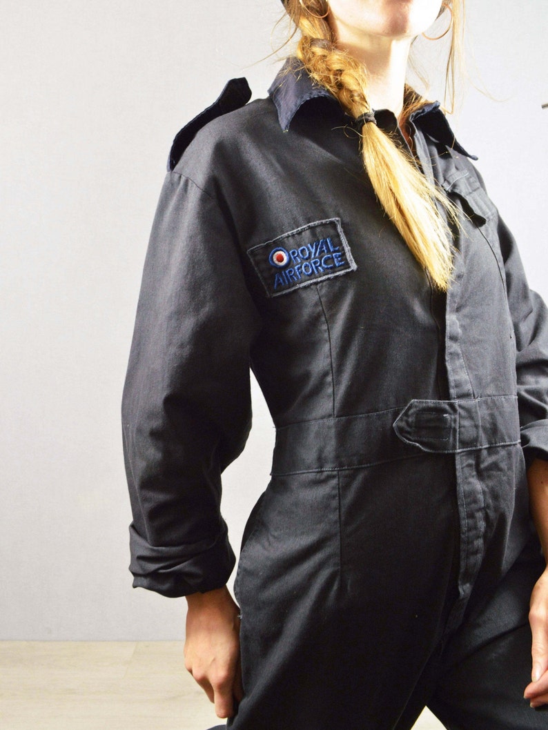 May include: Dark navy blue jumpsuit with a collared neckline and long sleeves. Features a belt at the waist and a patch on the chest that reads "ROYAL AIRFORCE" in blue and white lettering. The jumpsuit is made of a durable fabric.