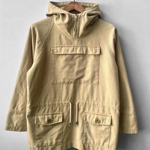 May include: A beige hooded jacket with a front zip, a large front pocket, and two lower pockets with button fastenings. The jacket features a drawstring at the waist and is hanging on a wooden hanger.