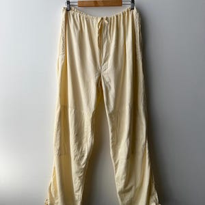 Peut inclure: Pantalon crème à cordon de serrage suspendu à un cintre en bois. Le pantalon a une coupe décontractée avec une taille à cordon et des liens aux chevilles. Le tissu semble léger et adapté aux climats chauds. Le pantalon est présenté sur un mur blanc uni.