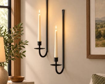 May include: Two black metal wall-mounted candle sconces with long, slender arms. Each sconce holds a single cream-colored taper candle. The candles are lit, emitting a warm glow against the wall. The sconces feature a simple, curved design.