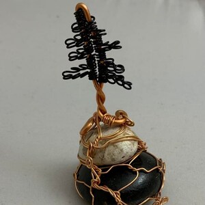 May include: A wire sculpture of a tree with black wire branches and a copper wire trunk. The tree is perched on top of two stacked stones, both wrapped in copper wire.
