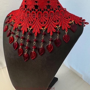 May include: A red lace collar necklace with leaf-shaped pendants. The collar is made of intricate red lace with a floral design. Red leaf-shaped pendants hang from the collar on gold chains. The necklace is displayed on a black mannequin.