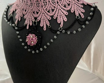 May include: A black and pink lace choker necklace with a black and pink flower pendant. The pendant is adorned with rhinestones. The necklace is on a black display stand. "WireWorkandWhatNot" is written in pink letters on the stand.