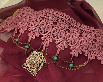 May include: A dusty rose lace choker featuring an intricate floral pattern. Attached to the lace are bronze-toned metal chains adorned with small turquoise-colored beads. A rectangular bronze-toned pendant with pink and turquoise accents hangs below.