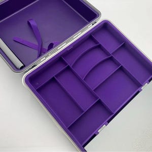 May include: A silver-toned hard-shell carrying case with a vibrant purple interior. The case features a deep open compartment with purple ribbon ties and a separate section with multiple rectangular and curved dividers for organization.