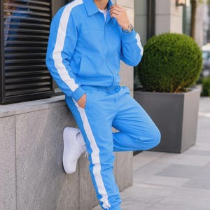 Könnte beinhalten: Ein leuchtend blauer Trainingsanzug mit einer Jacke mit Reißverschluss und Kragen sowie einer passenden Hose, beide mit einem markanten weißen vertikalen Streifen an den Ärmeln und Beinen. Dazu ein blauer Fischerhut und weiße Sneaker.