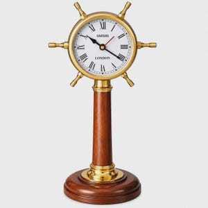 May include: A table clock featuring a brass ship's wheel frame with a white face, black Roman numerals, and black hands. The text SMISSS and LONDON is printed on the face. It stands on a polished wooden pedestal base with brass accents.