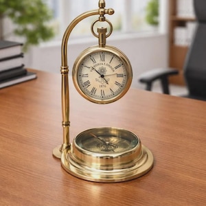 May include: A brass-colored desk clock and compass set featuring a circular clock face with Roman numerals, the text Victoria London 1876, and an anchor icon. The clock hangs from a curved stand above a circular magnetic compass base.
