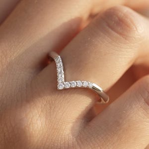 May include: A silver-toned chevron-shaped ring featuring a row of small, clear, round-cut stones set along the V-shaped band. The metal band has a polished, reflective finish, and the stones are arranged symmetrically to follow the angular design.