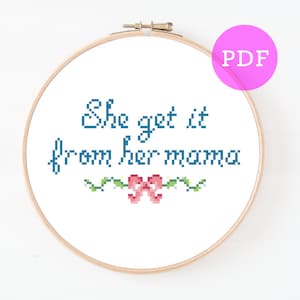 以下が含まれることがあります： 白い刺繍枠に「She get it from her mama」という文字が青い糸で刺繍されています。文字の下には、小さなピンクのリボンと緑の葉が飾られています。