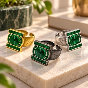 Könnte beinhalten: Drei verstellbare Ringe mit einem kreisförmigen grünen Emblem und einem zentralen Spiralmuster. Die Ringe sind in Gold-, Dunkelgrau- und Silbertönen gehalten und weisen jeweils eine texturierte grüne Glitzereinlage auf dem Emblem auf.
