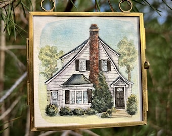 May include: A square watercolour painting of a two-storey house with white siding, a prominent brick chimney, and dark shutters, encased in a thin gold metal frame with a hanging chain. The house features a front door, multiple windows, and green trees.